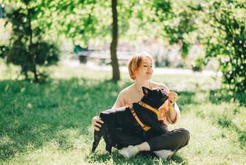 Black french Bulldog Puppy cuddling up with owner girl laying on lawn in park outdoors summer spring day kissing hugging petting. Happy woman with dog outside