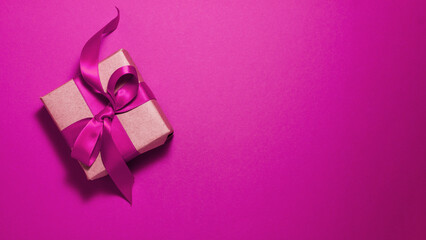Brown paper gift box with a dark pink satin ribbon bow on pink background on the left with copy space. Flat lay Valentines day father mother day and presents concept.