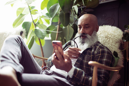 Mature man with beard using smart phone in armchair