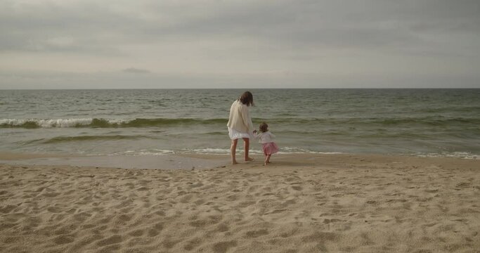 Young mother and baby daughter spend time on the beach together. Mom and toddler cute little girl walk by the sea alone holding hands. Motherhood, maternity, single mother, family concept.