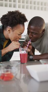 Vertical video of happy african american couple drinking juice from one glass with straws