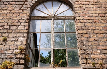 Alte rot-braune Backsteinfassade mit Rundbogenfenster und eingeschlagenen Glasscheiben vor Baum und blauem Himmel bei Sonne am Nachmittag im Sommer