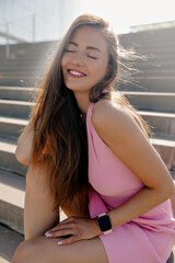 lovely adorable lady in pink dress with long loose hair smiling with closed eyes and enjoying sunlight on stairs in the city