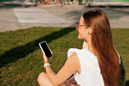 Close up portrait from back of young woman with long hair wearing white dress sitting on green lawn in headphones holding smartphone and resting in sunny morning