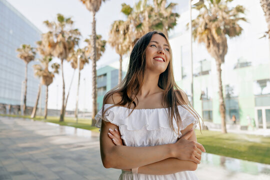 Spectacular pretty woman with wonderful smile wearing white summer dress posing on background of palms in Spanish city. Caucasian fair-skinned smilies and look away 