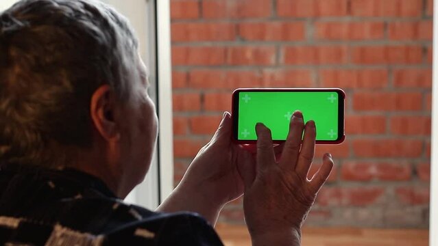 senior woman holding cell phone horizontally and press virtual keys of green touch screen display, for mock-ups showing app development or graphic design. Place mobile app logo on green chroma key tou