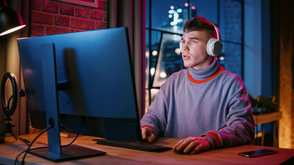 Young Handsome Man in Headphones Using Computer in Stylish Loft Apartment in the Evening. Creative Male Smiling, Working from Home, Browsing Videos on Social Media. Urban City View from Big Window.