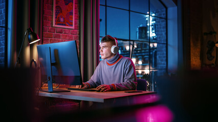 Young Handsome Man in Headphones Using Computer in Stylish Loft Apartment in the Evening. Creative Male Smiling, Working from Home, Browsing Videos on Social Media. Urban City View from Big Window. - obrazy, fototapety, plakaty