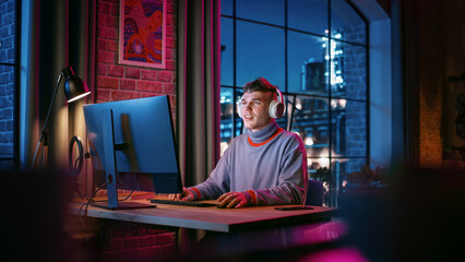 Young Handsome Man in Headphones Using Computer in Stylish Loft Apartment at Night. Creative Male Working from Home, Browsing Internet and Social Media. Urban City View from Big Window. © Gorodenkoff