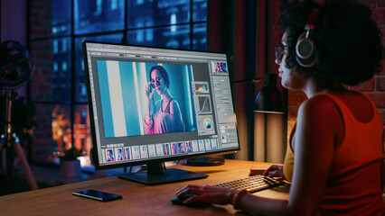 Young Creative Female in Headphones Using Computer in Stylish Loft Apartment in the Evening. Graphic Designer Working from Home, Editing Fashion Photo for Online Store. Urban View from Big Window.