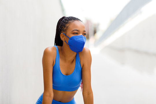 A Black Woman Runner Stands In A Mask From The Coronavirus, Bending Over And Taking A Breath After A Run