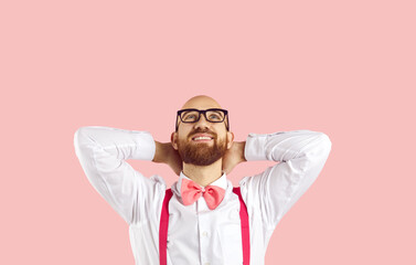 Young happy man holds his hands behind his head and dreamily looks up on pastel pink background. Caucasian handsome stylish man in white shirt, bow tie and suspenders dreams or imagines. Copy space.