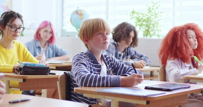 Focused male student studying at school with classmates. Thoughtful schoolboy in casual wear writing in notebook during lesson in bright classroom.