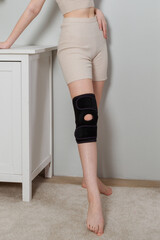 Black knee support brace on a woman leg. girl in an orthosis in the interior of the house....