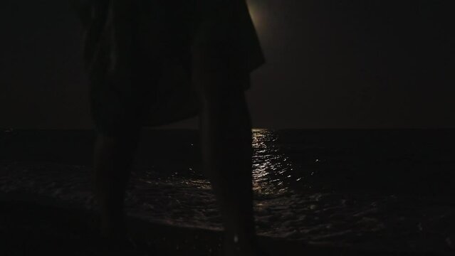 The girl walks along the sea beach at night by the light of the moon. Moonlight on the water, waves.