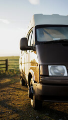 Fototapeta na wymiar Shot of a brown and white retro fashioned camper van laying near the wooden fence of a green field during the sun rise moments of the road trip.