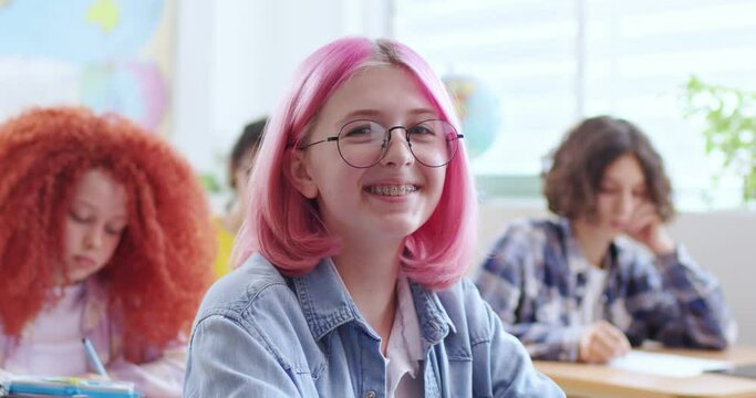 Smiling pink haired girl in glasses sitting at desk in classroom and looking at camera. Portrait of positive female student studying with male and female classmates on background.