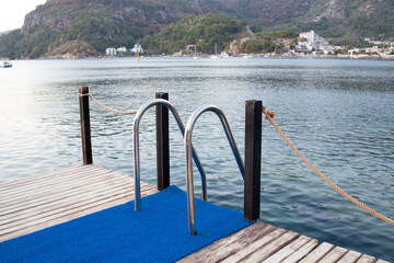 Fototapeta na wymiar Metal handrails and ladder to descend into the water on a wooden pier of a sea bay - a place for swimming. Wooden pier with ladder for comfortable entrance in the sea.