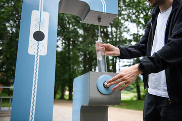Man refilling his water bottle at the city. Free public water bottle refill station. Sustainable and green city. Male in black coat. Tap water to reduce plastic bottle usage. Drinking water dispenser