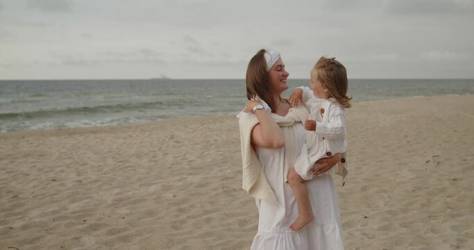 Young mother and baby daughter have fun spending time on the beach together. Mom holds her toddler cute little girl on hands by the sea kissing. Motherhood, maternity, single mother, family concept.