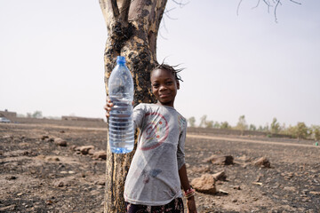 Beautiful little African girl standing in a dry stony field holding up an empty plastic bottle...
