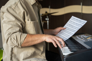 Young man checks music sheet and plays notes on synthesizer. Drum-kits stand in specifically equipped music studio close side view