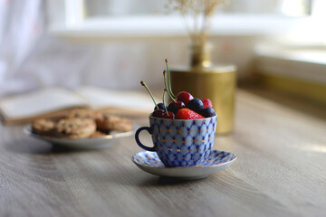 Plate of chocolate chip cookies, cup filled with strawberries, blueberries and cherries, open book and vase with gypsophila flowers on the table. Selective focus.
