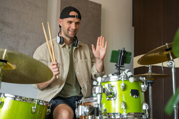 Young man greets followers recording online drum lesson on mobile phone and plays drums in equipped music studio. Guy explains nuances of drums playing technique