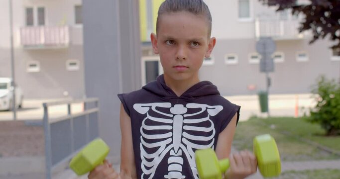 A teenager raises and lowers dumbbells in his hands in the courtyard house. He has a pattern of bones on his clothes. Hand training, increase the strength of the child. CZ, Kladno, Polska, 4.7.22