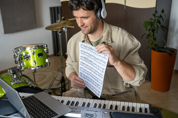 Man gives piano online lesson via laptop demonstrating notes on music sheet in specifically equipped musical studio with drum-kits on background