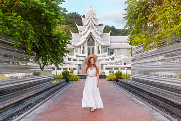 Traveler woman in white dress walking in front of a beautiful white temple in thai style. Travel and vacation in Asia. Young woman in hat near beautiful buddhist temple on Phuket Island in Thailand - obrazy, fototapety, plakaty