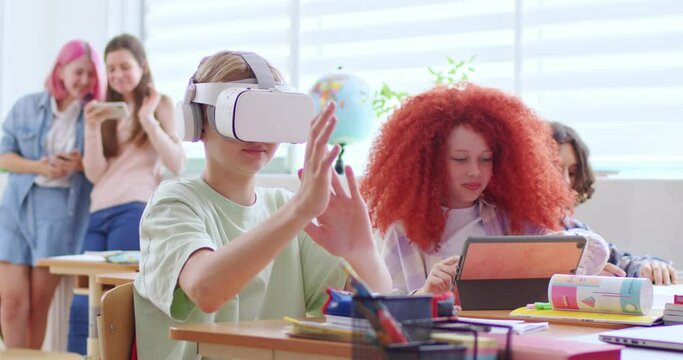 Smiling girl with red hairstyle setting up different programs on tablet for her male classmate in vr headset. Excited boy playing games wearing goggles in classroom during break.
