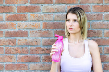 Fototapeta na wymiar Close up portrait of an attractive young sporty blonde caucasian woman wearing pink sports attire drinking water from a bottle High quality photo