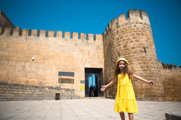 A tourist girl in a yellow hat and sundress walks along the street of the old town made of stone with a fortress. Sightseeing tour. The child got lost, searching for parents