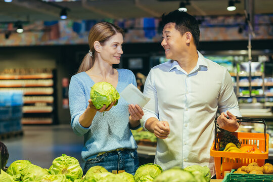 Happy Mixed Race Asian Couple Family Man And Woman Choosing Vegetables In Grocery Store Supermarket. Make A Purchase Daily Shopping. Customer Select Product Pick An Cabbage In Eco Bag In Food Market