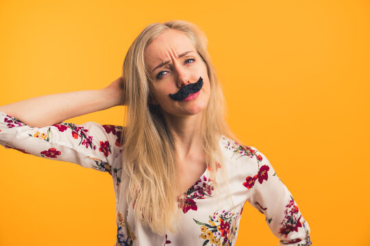 Funny Long-haired Blond Caucasian Woman In Her Mid 20s With Fake Black Moustache On Her Face. Confused Facial Expression. Medium Closeup Studio Shot Over Orange Background. High Quality Photo