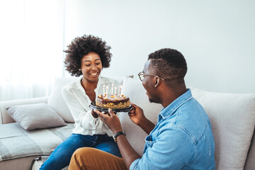 Happy African American couple celebrating anniversary or wife birthday at home. Portrait of happy woman blowing out candles on birthday cake standing with husband together in Living room