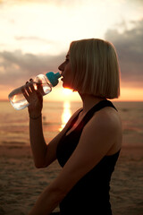 fitness, sport, and healthy lifestyle concept - woman drinking water from bottle on beach over...