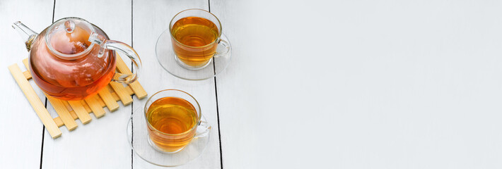 Tea in glass teapot and two cups on a wooden white background. Teapot with green or black tea with reflection in glass in the country. Banner