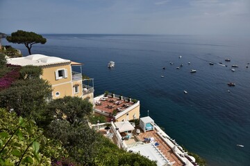 Fototapeta na wymiar Iconic view of ritzy terraces full of sun loungers against Mediterranean stone pine, bougainvillea flora and white yachts in azure ocean waters of the Gulf of Salerno near Positano, Italy
