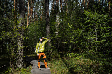 A man in the forest stands on a trail of boards, a wooden road, an ecological trail, a traveler explores the countryside.