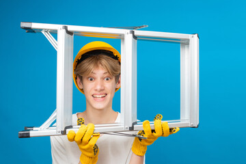 Happy teen in white t-shirt and gloves with yellow helmet in head, holding ladder in hands standing...