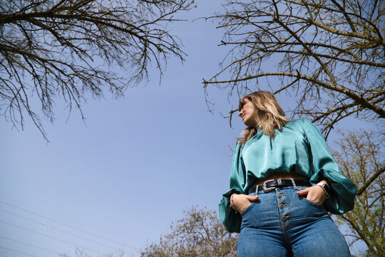 Young, Beautiful Blonde Woman Is Looking Down At The Camera From Above, In The Background You Can See The Blue Sky And Branches Of Trees On A Sunny Day. The Woman Is Happy. Concept Expressions.