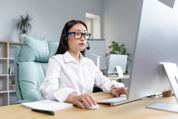 Obraz na płótnie Canvas Online distance learning. Young beautiful Asian female teacher in headphones and glasses sitting at a table at a computer teaching a class lesson online.