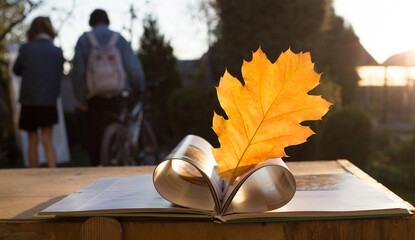 Education concept. Hello, Autumn. In the foreground is an open book with an orange oak leaf between open heart-shaped pages. Back to school. Couple of students in the background out of focus