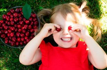 Close-up of smiling caucasian girl  in a red T-shirt with cherries on her closed eyes lies on the grass in summer. Child's face in the rays of the sun