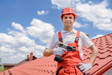 young smiling man worker master in red overall and helmet is fixing the metal tile roof with electric screwdriver.