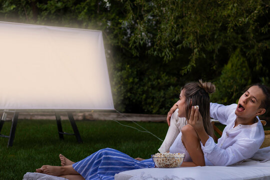 Young Couple In The Park Watching A Summer Movie. Boyfriend Stealing Opcorns From Behind To His Girlfriend