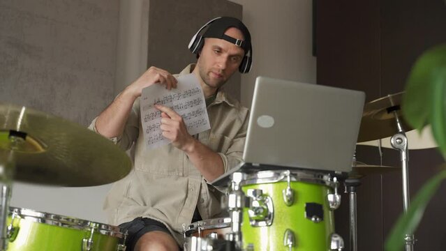 Young man records drum lesson via laptop and demonstrates notes on music sheet in specifically equipped music studio. Guy explains details showing notes