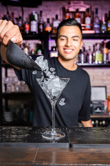 young and smiling Latin American waiter throws ice cubes to a glass of cocktail